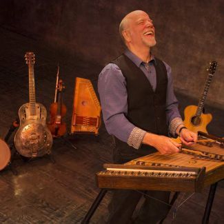 musician plays hammered dulcimer in front of a row of guitars