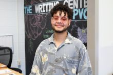 A smiling person with curly hair wearing pink-tinted round glasses and a floral shirt, standing in an office-like setting with a sign reading "THE WRITING CENTER @PVCC" in the background.